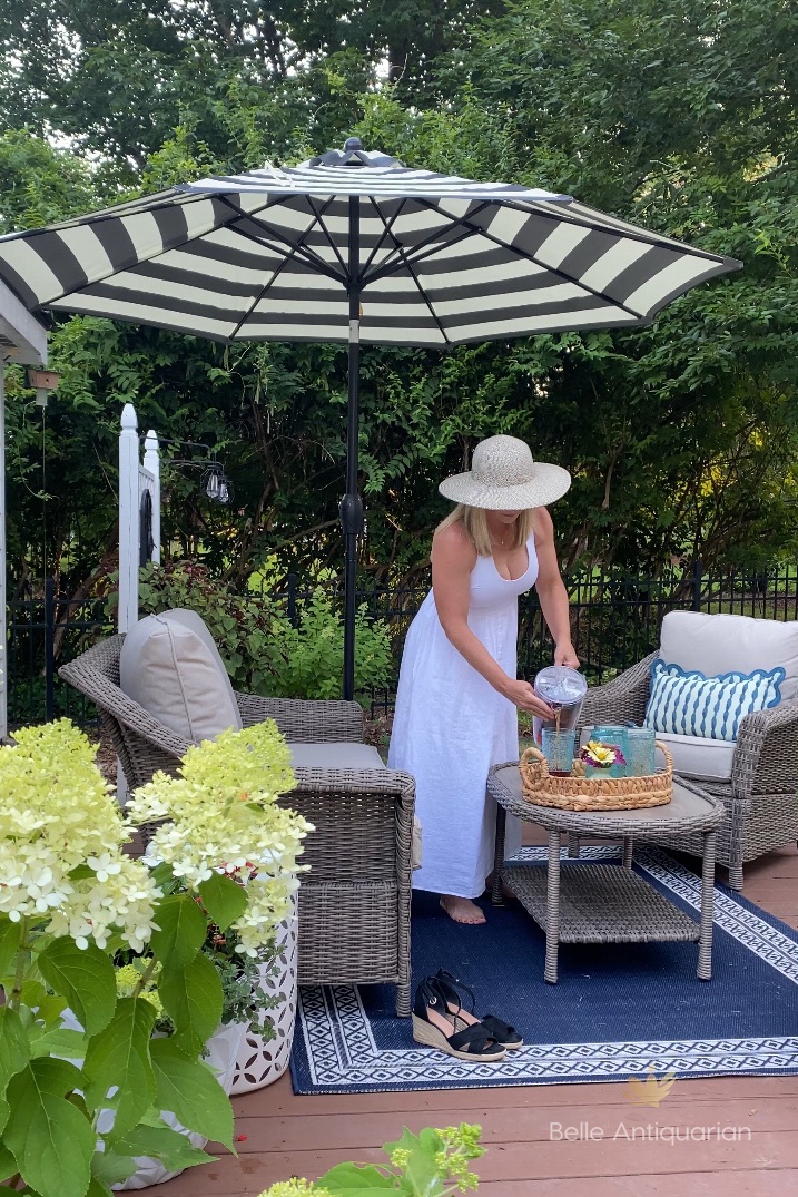 A southern garden at golden hour. A woman wearing a white linen dress and a straw hat pours a glass of sweet tea.