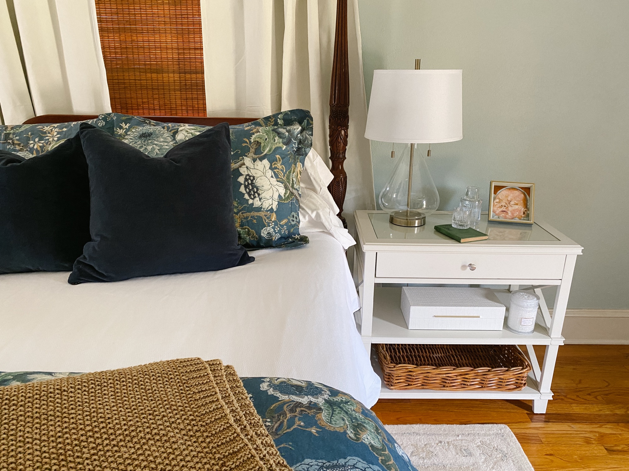 Feminine nightstand with lamp, carafe, small art, storage boxes, candle, and basket.
