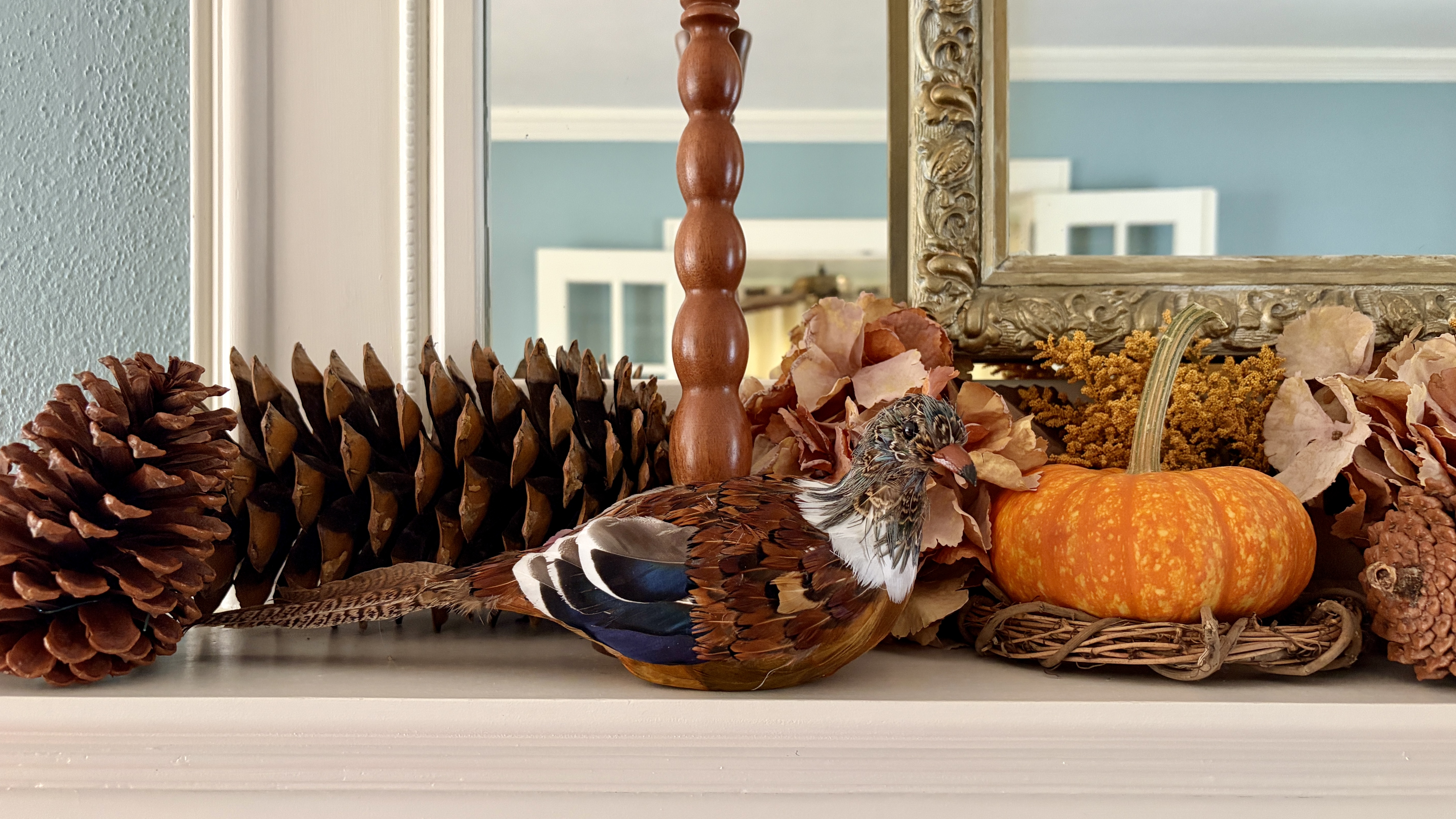 Fall decorating on a mantel with a pheasant figurine, pumpkin on a grapevine wreath, large pinecones, wooden candlesticks, and a gilt mirror.
